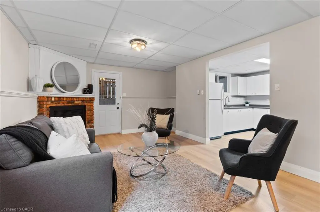 Living room with gray sofa, black chairs, and brick fireplace. A glass table sits on a gray rug. A white kitchen is visible through an opening.