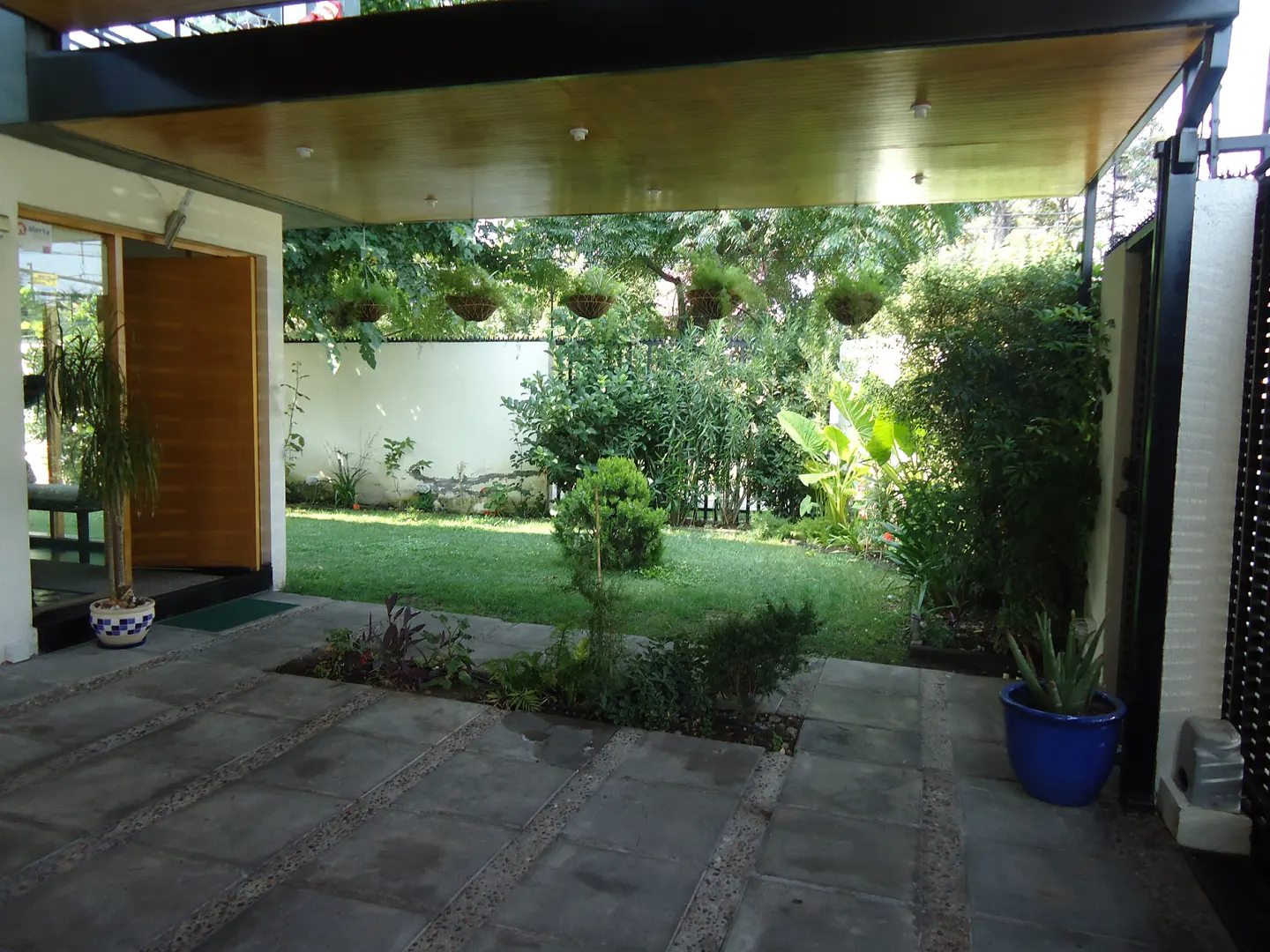 Covered patio with gray stone tiles leads to a green lawn and garden. Hanging plants and a blue pot add color.