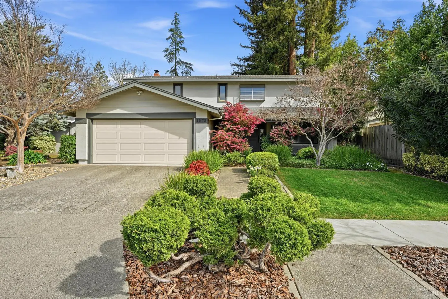 Two-story house with a beige exterior, a two-car garage, and a well-manicured lawn with green bushes and a red-leafed tree.