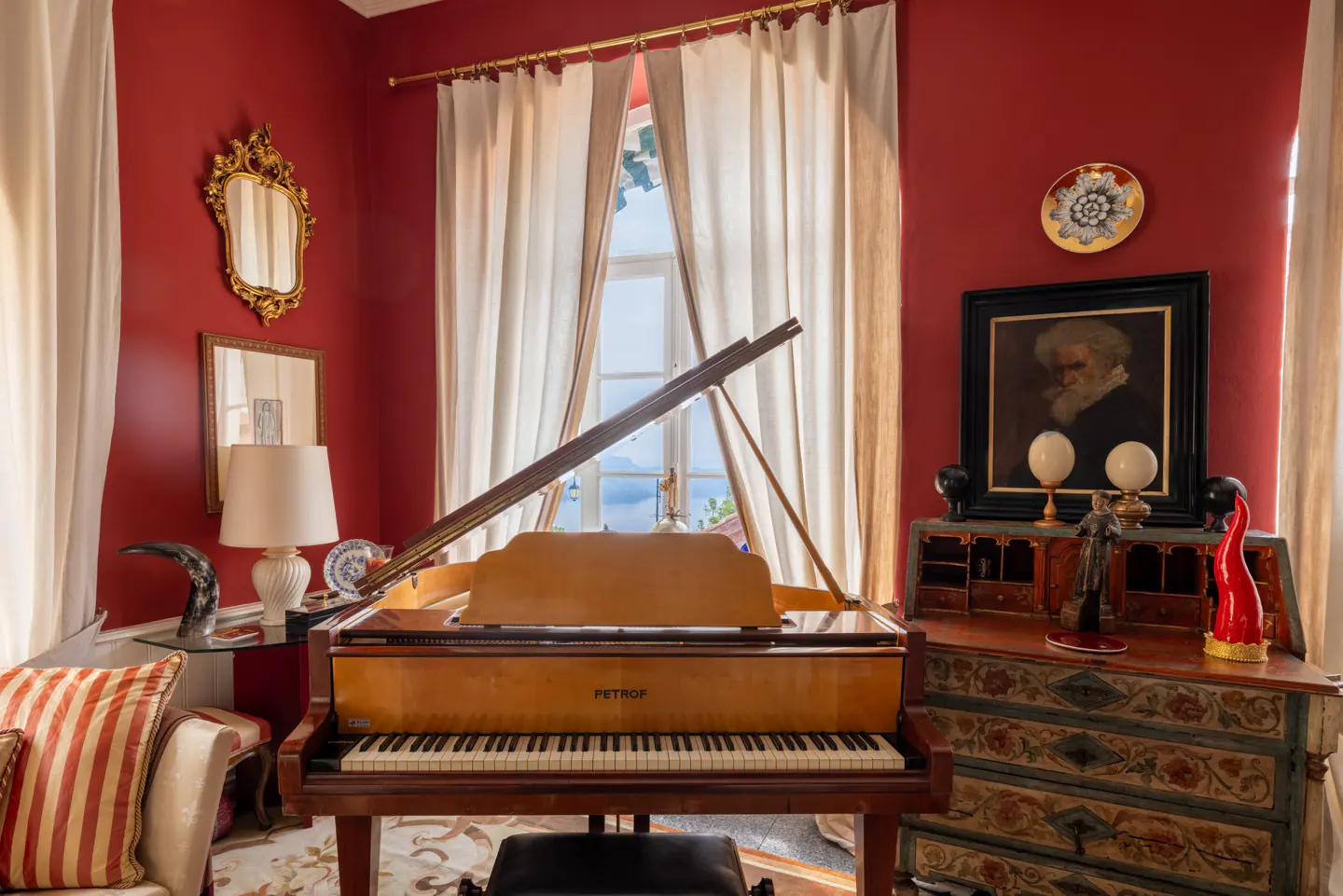 Interior view of a red room with a grand piano, antique desk, and white curtains framing a window with a sea view.