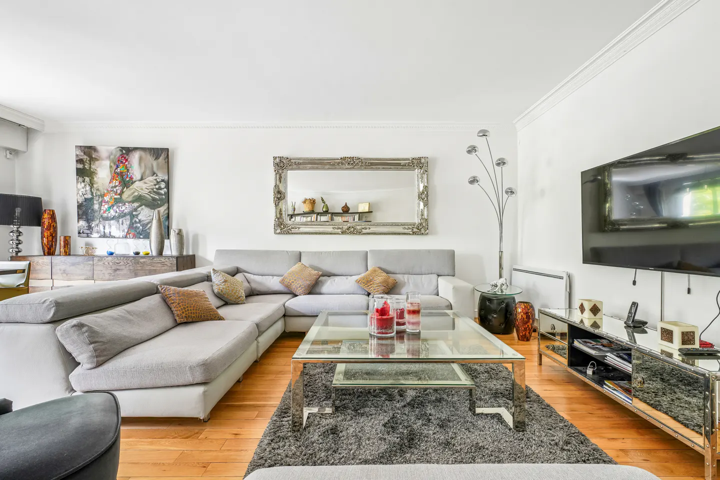 Bright living room with a gray sectional sofa, glass coffee table, and a large mirror on the white wall. A flat-screen TV is mounted on the wall.