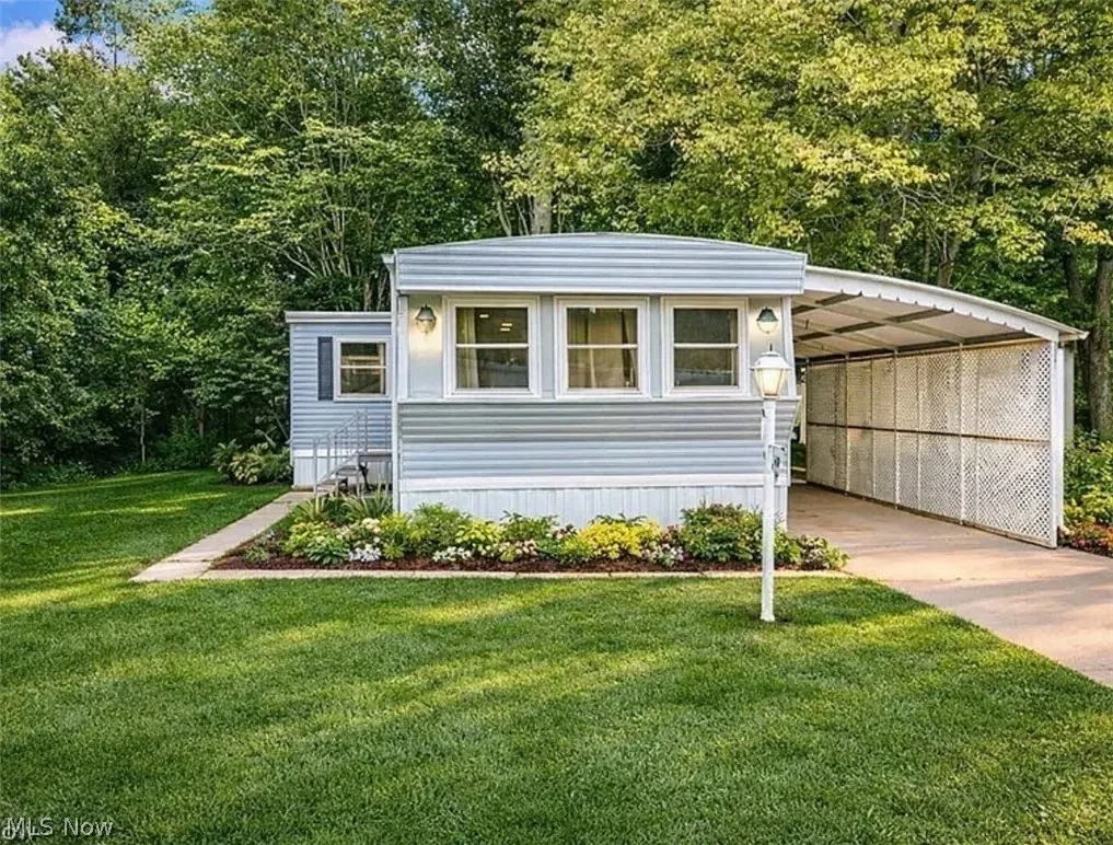 A light blue mobile home with white trim, a carport, and a green lawn. Trees are in the background.