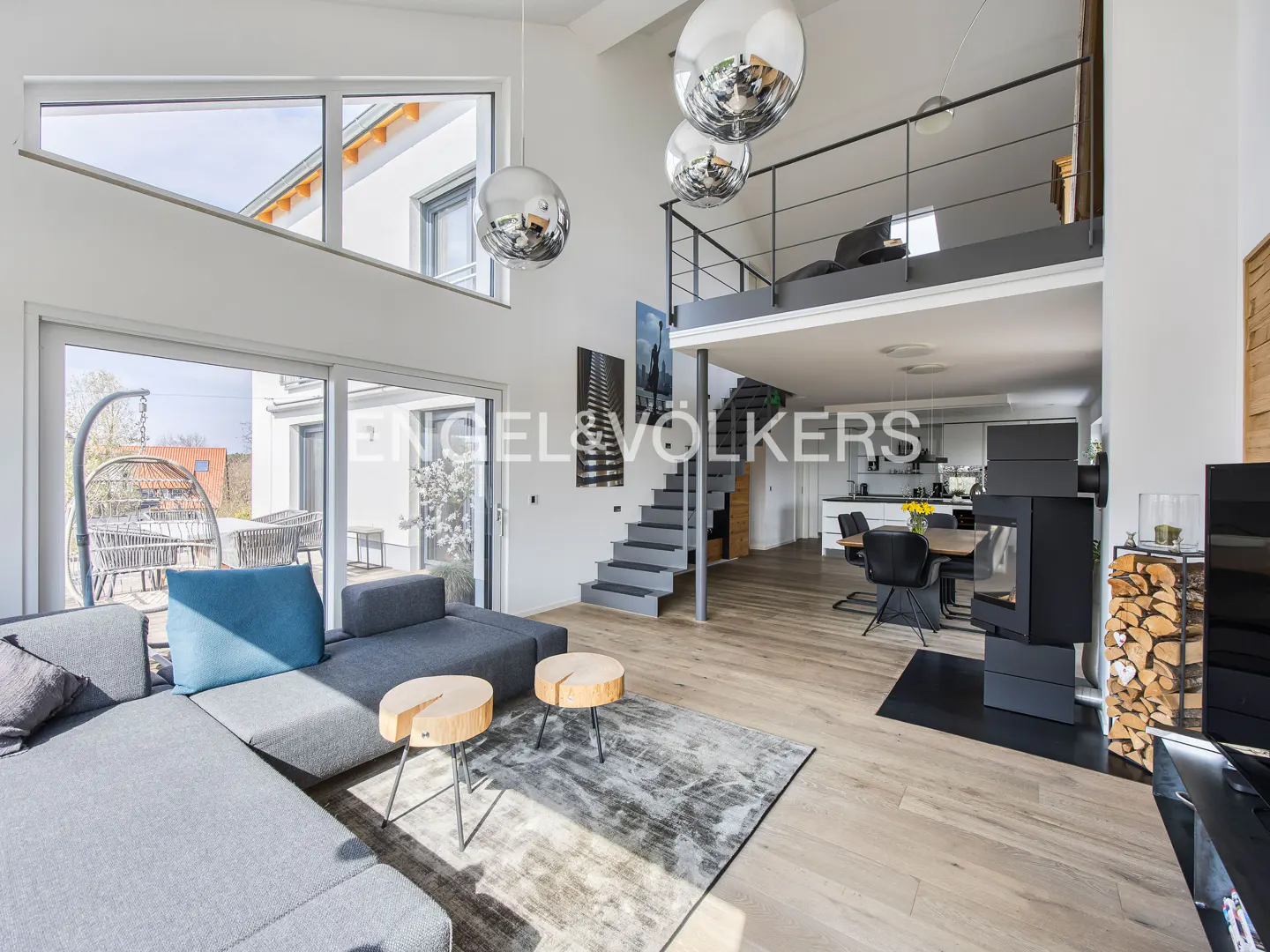 Bright, modern living room with gray sectional, wood tables, and a fireplace. Stairs lead to a loft area. Large windows offer outdoor views.