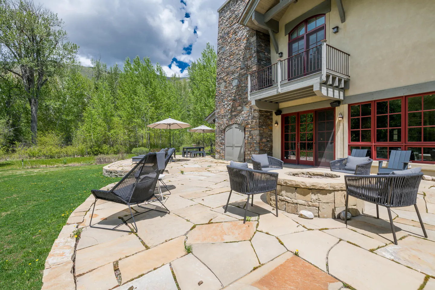 Outdoor patio with stone fire pit and black woven chairs. A stone building with red framed windows and a balcony is in the background.
