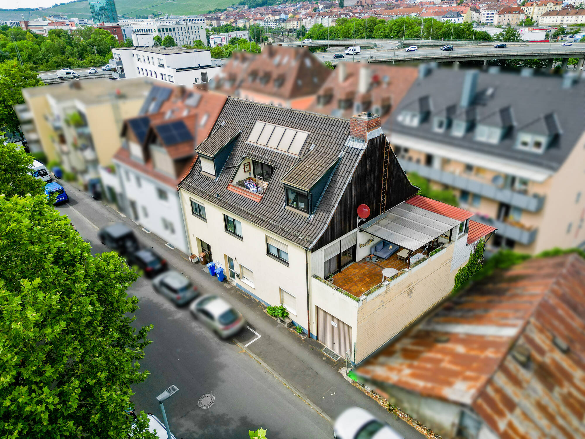 Aerial view of a house with a dark roof, rooftop terrace, and garage, located on a street near a highway and city buildings.