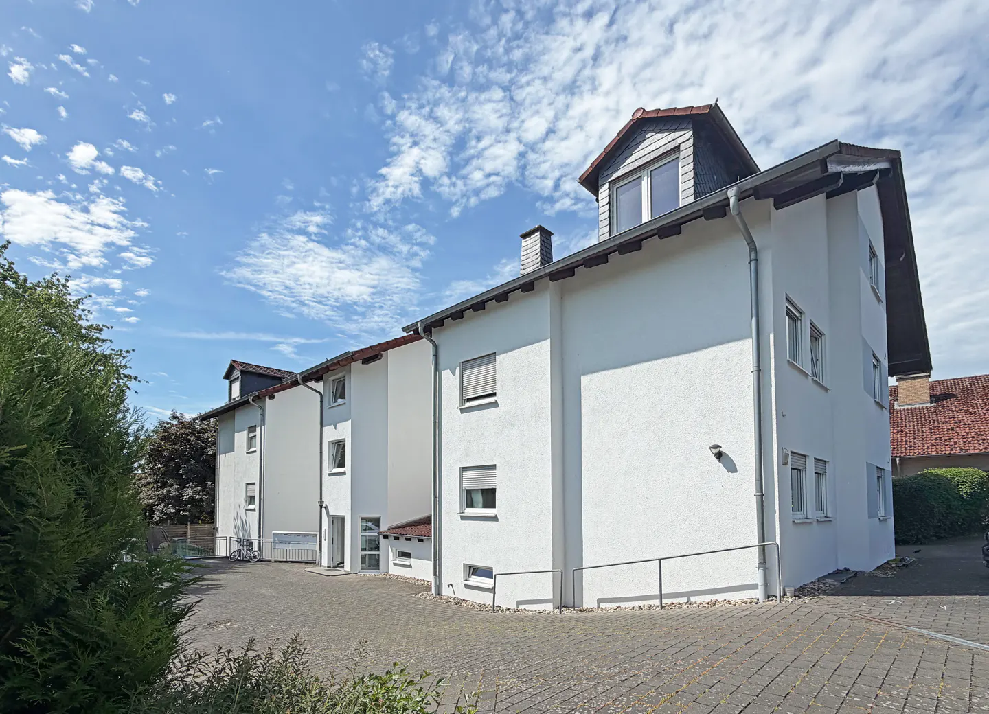 Exterior view of a white multi-story apartment building with a paved courtyard under a bright blue sky with clouds.