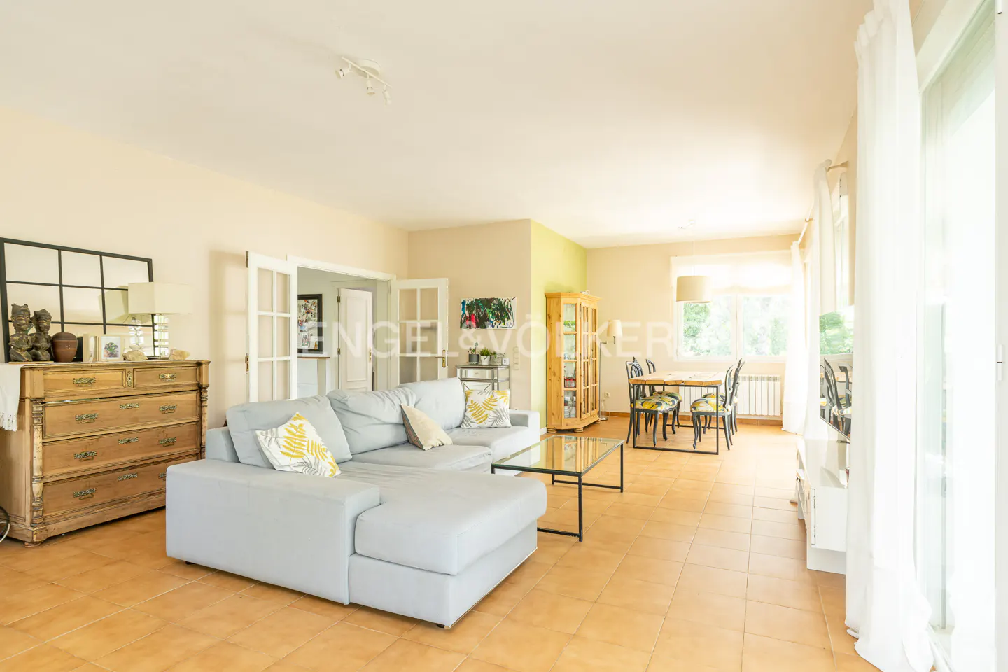 Bright living room with a gray sectional sofa, glass coffee table, and dining set. Natural light fills the space with tan tile flooring.