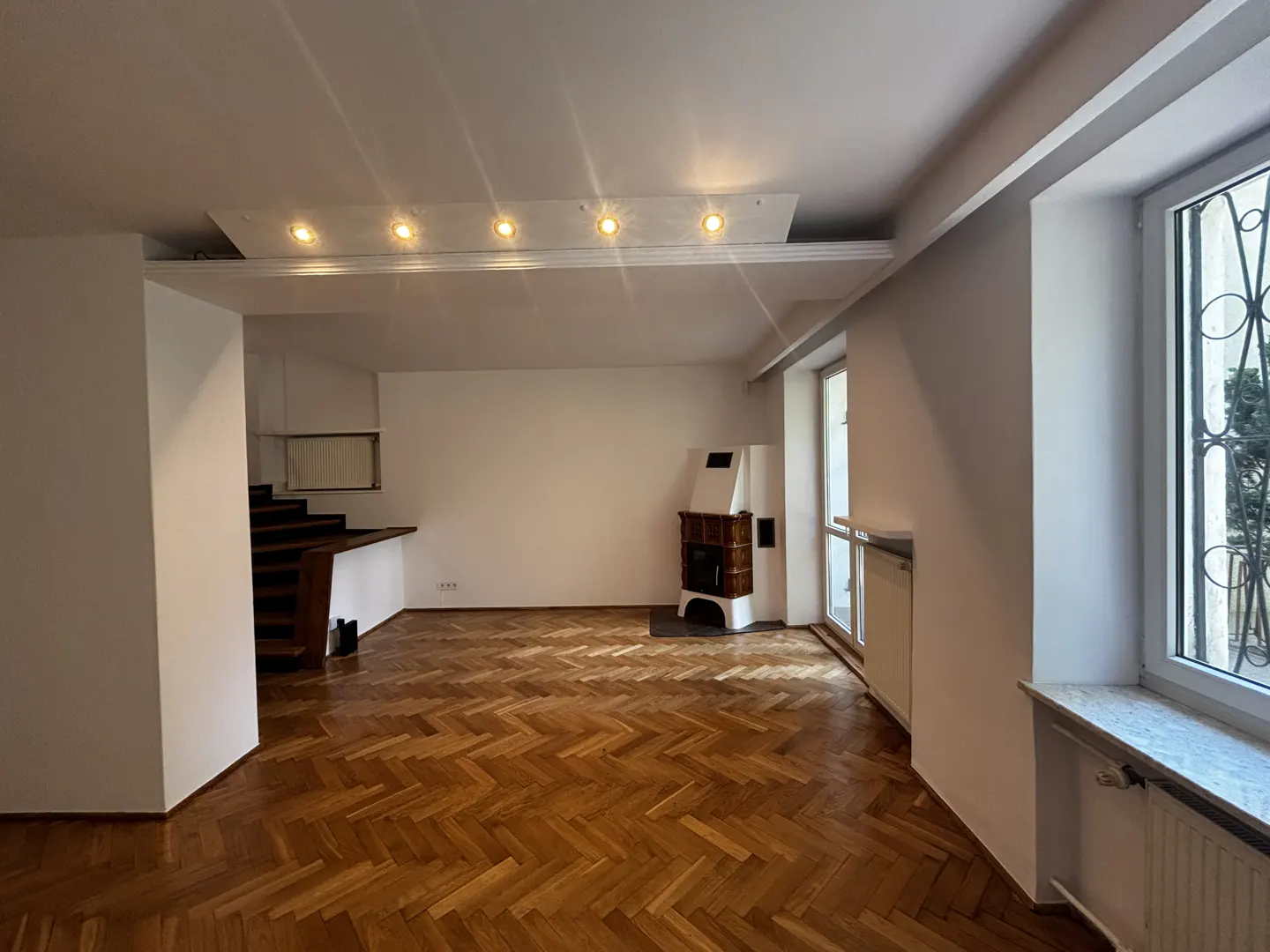Bright, empty living room with herringbone wood floors, white walls, and a decorative fireplace. Stairs are visible on the left.