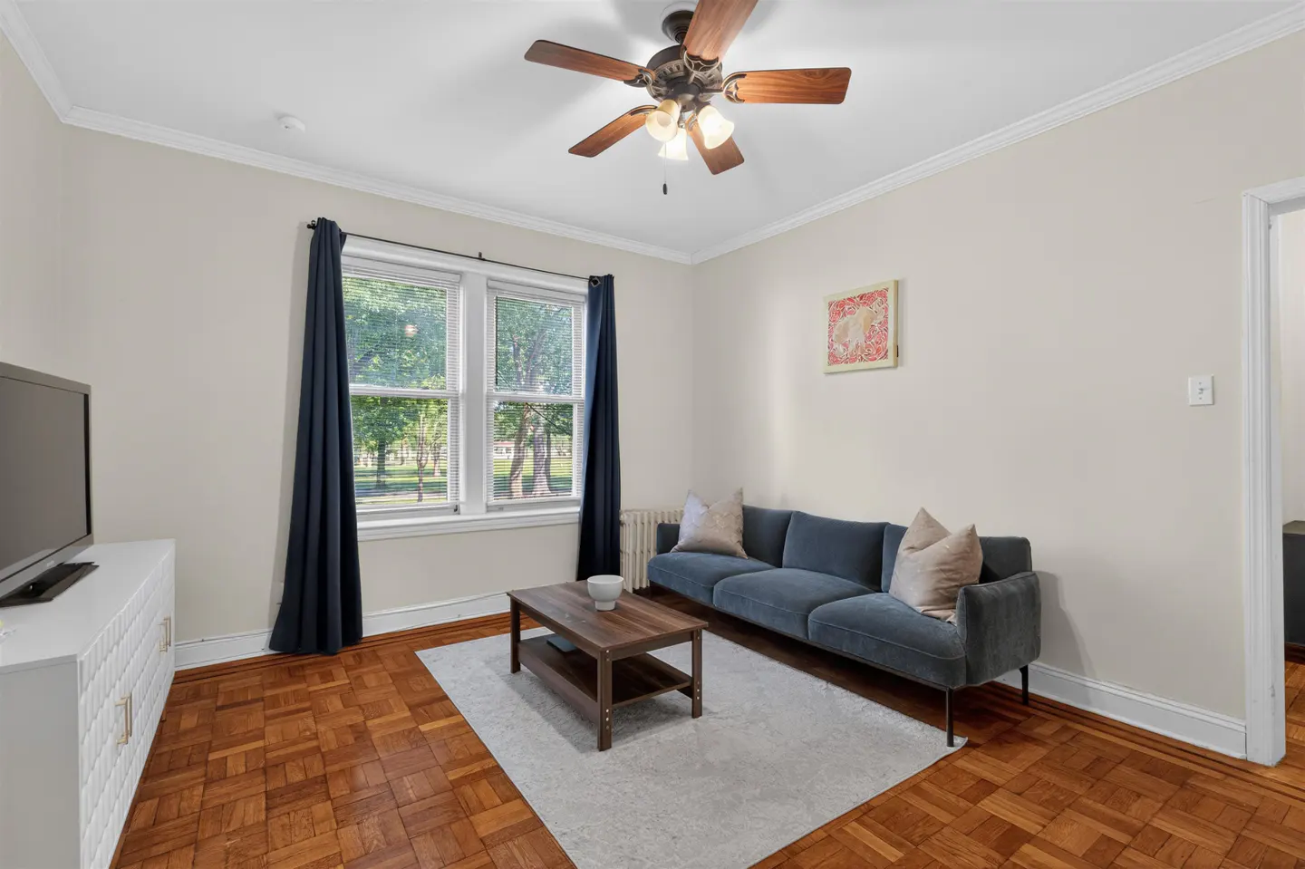 Living room with blue sofa, wooden coffee table, parquet floors, ceiling fan, and windows with dark curtains.