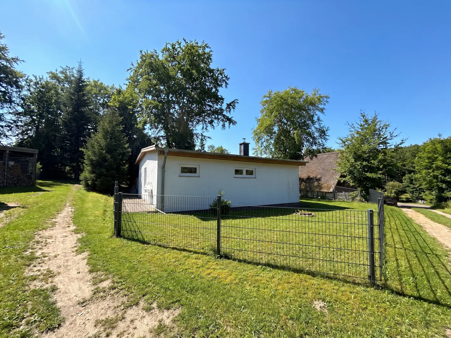 Exterior view of a white, one-story house with a black chimney, surrounded by a green lawn and a wire fence. Trees and a blue sky are in the background.