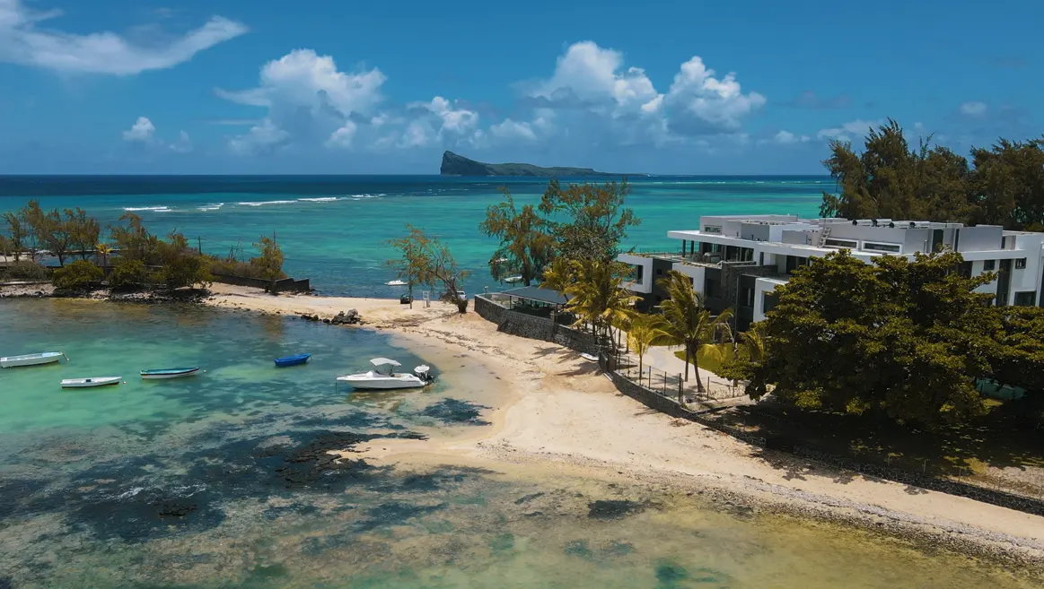 Aerial view of a white modern beachfront property with boats on turquoise water and a distant island under a blue sky.
