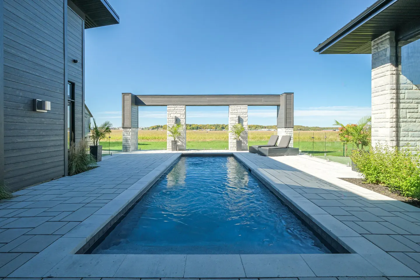 Rectangular pool with blue water, stone patio, and pergola overlooking a field on a sunny day. Two lounge chairs are visible.