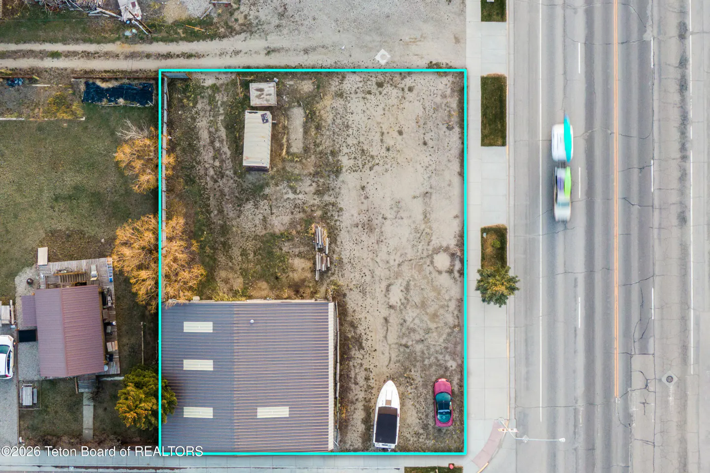 Aerial view of a vacant lot outlined in blue, next to a road with blurred cars, a building, and some trees.
