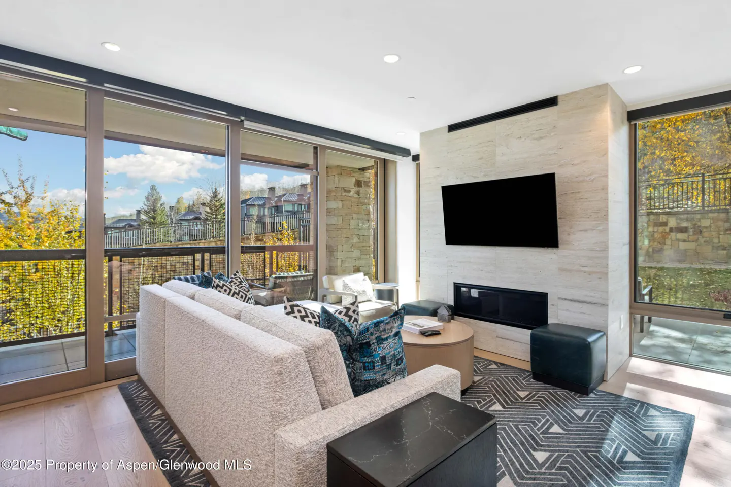Living room with a beige sofa, patterned pillows, and a black TV above a fireplace. Large windows offer a view of trees and buildings.