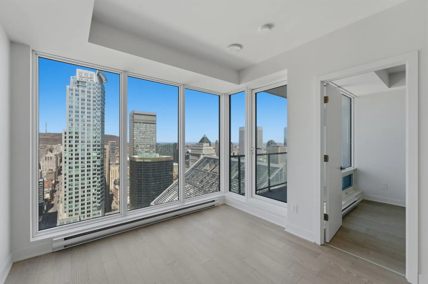 Bright, empty room with light wood floors and white walls. Large windows offer a city view. An open doorway leads to another room.