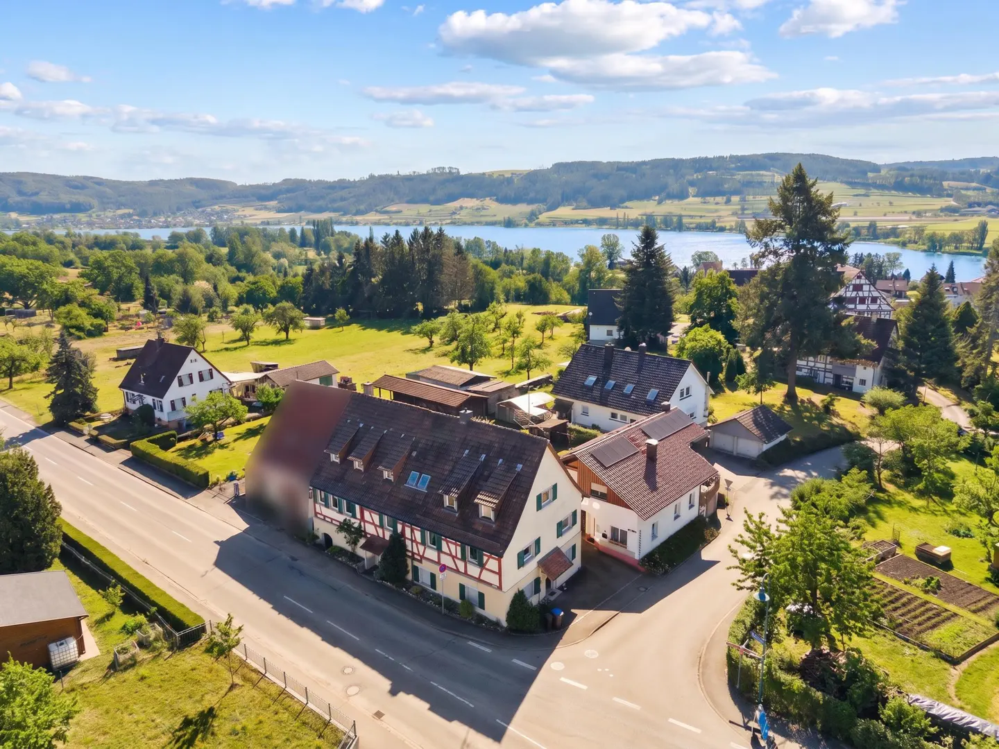 Aerial view of a white, half-timbered house with a red roof, surrounded by green trees and a lake in the background.