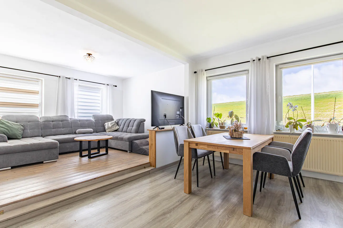 Bright, modern living room with a gray sectional sofa, wood table, and dining set near windows with a green field view.