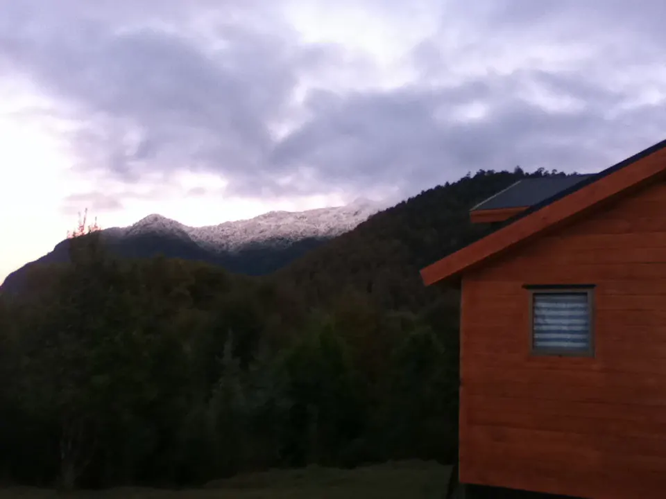 A wood cabin with a window sits before a mountain range with snow-capped peaks under a cloudy sky.