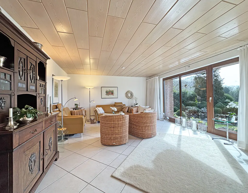 A bright living room with a wood-paneled ceiling, wicker chairs, and a large window overlooking a lush garden.