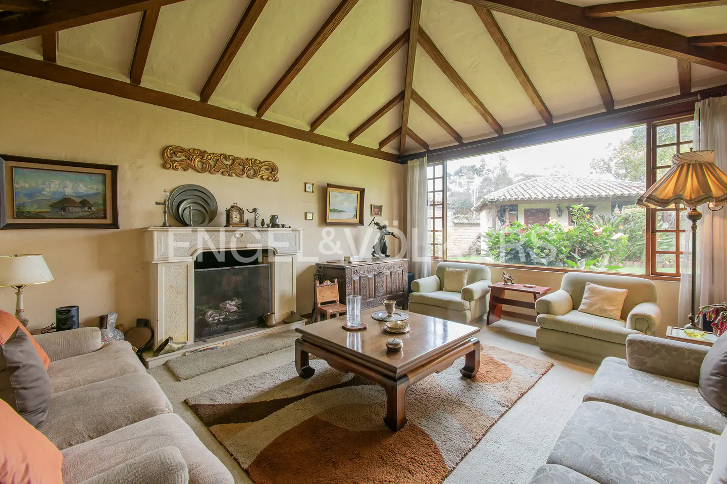 Living room with beige sofas, a fireplace, and a large window overlooking a garden with a house.