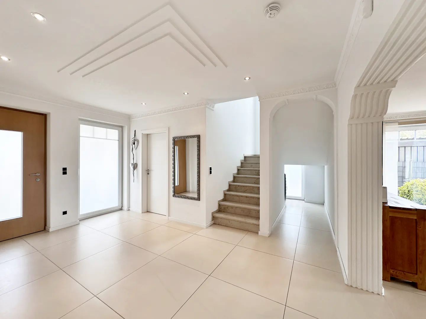 Bright foyer with beige tile floor, white walls, and stairs. A brown door is on the left, and a mirror hangs on the wall.
