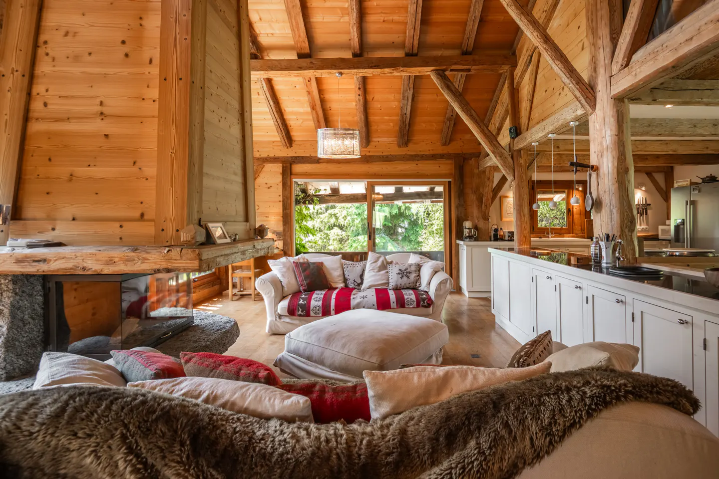 A cozy living room with wood walls, beams, and a stone fireplace. A white sofa with red pillows faces a large window with green trees outside.
