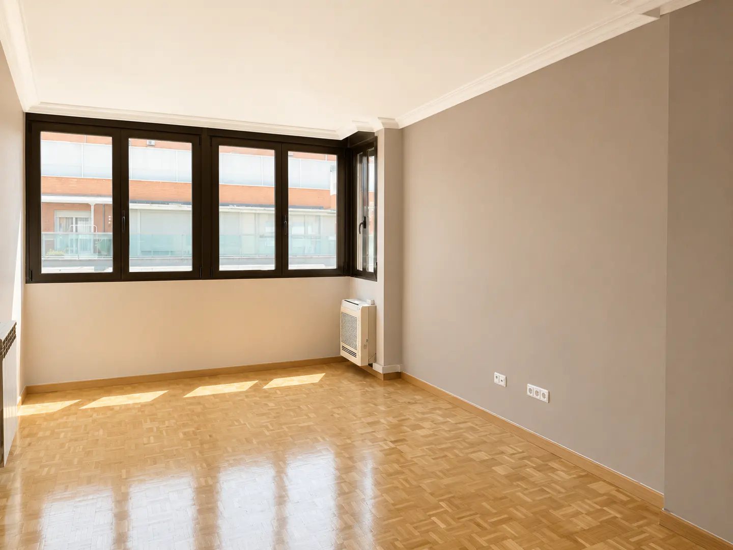 Empty room with light wood parquet flooring, beige walls, and large windows overlooking an apartment building.