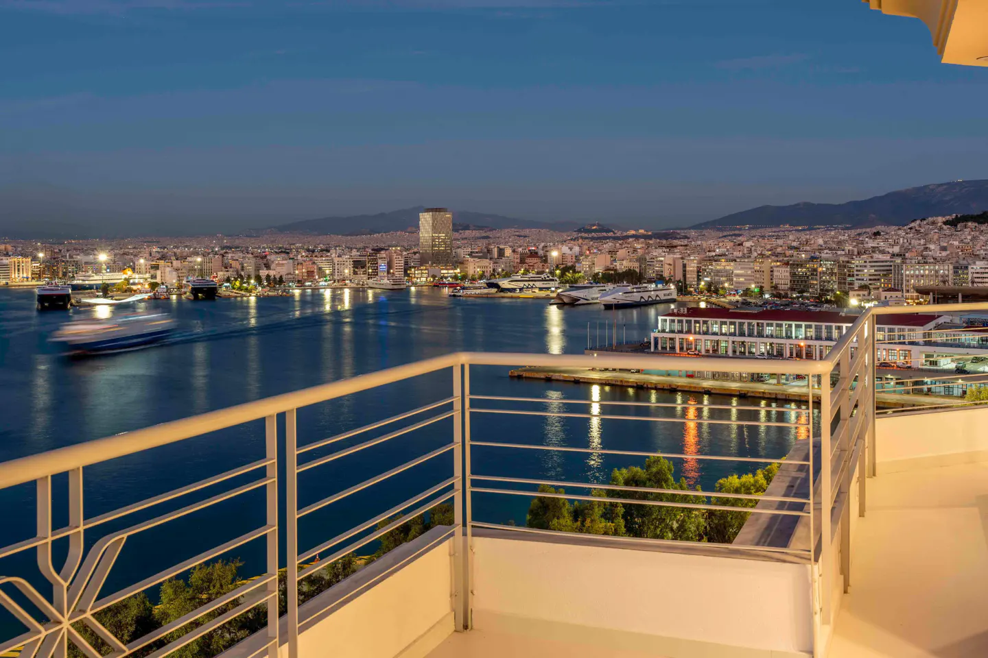 View from a balcony overlooking a harbor at dusk. White railings frame the scene, with city lights reflecting on the water.
