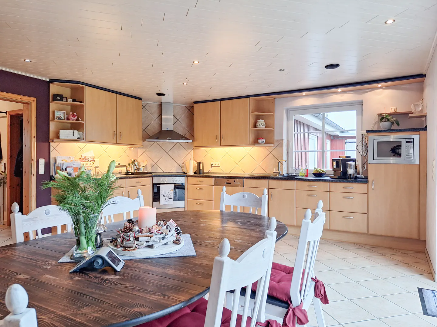 A bright kitchen with light wood cabinets, stainless steel appliances, and a dark wood table with white chairs and red cushions.