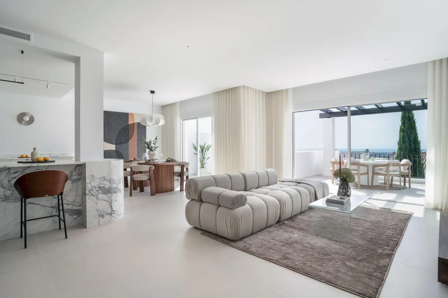 Bright, modern living space with a gray sofa, glass table, and marble kitchen island. Dining area and outdoor patio visible.