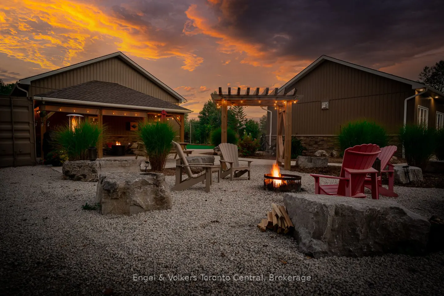 Outdoor patio with fire pit, Adirondack chairs, and pergola at dusk. Buildings flank the space, with a dramatic orange and purple sky above.