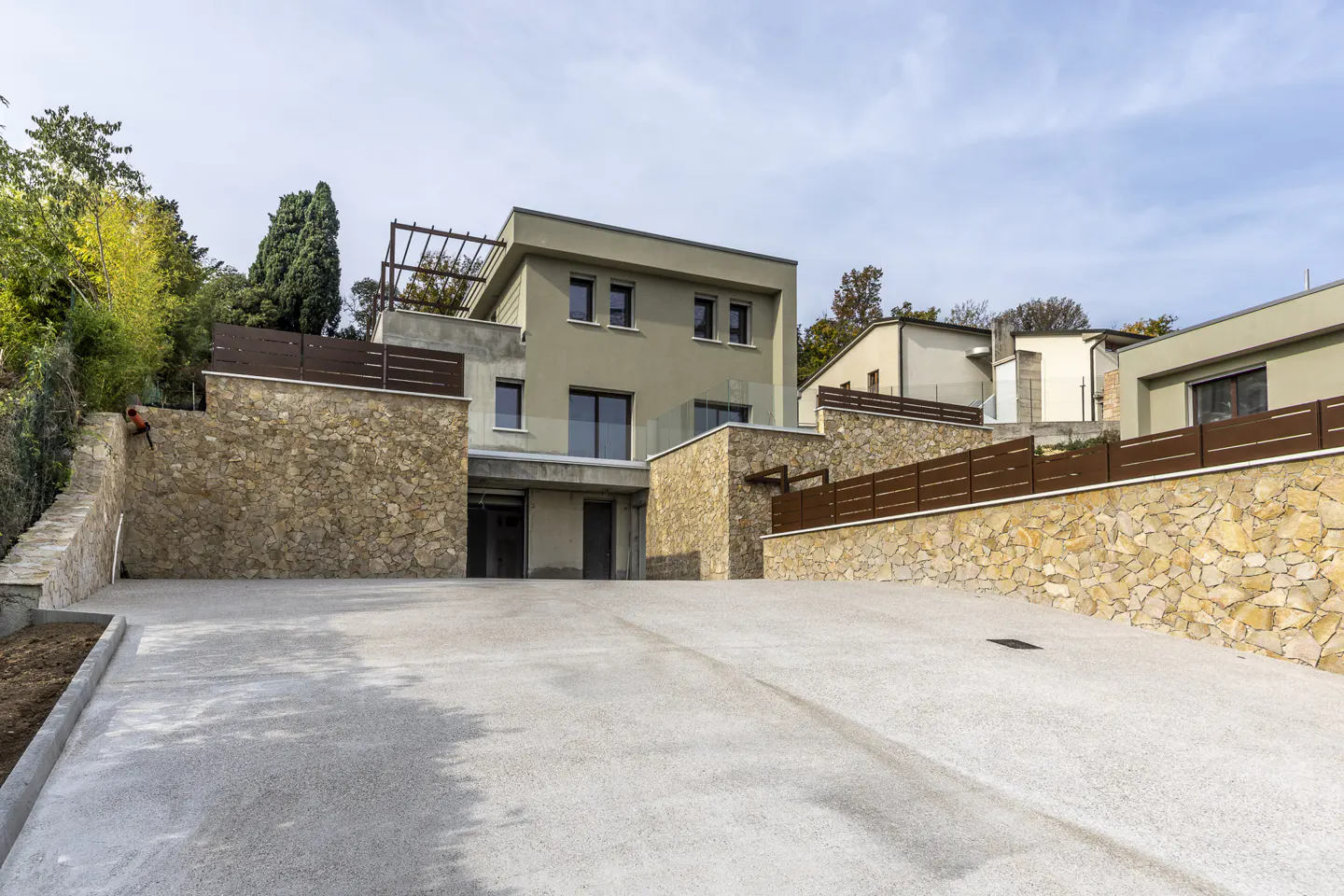 Modern two-story house with a flat roof and a rooftop pergola. The house is light green with stone walls and a concrete driveway.