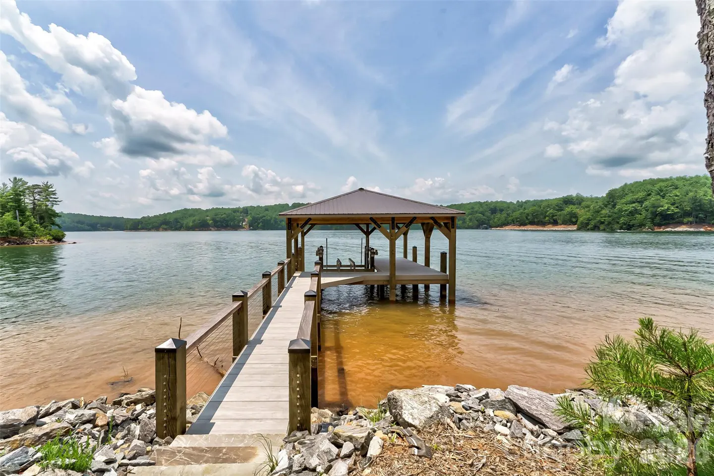 Lake view with a wooden dock and gazebo extending into the water. Green trees line the shore under a partly cloudy sky.