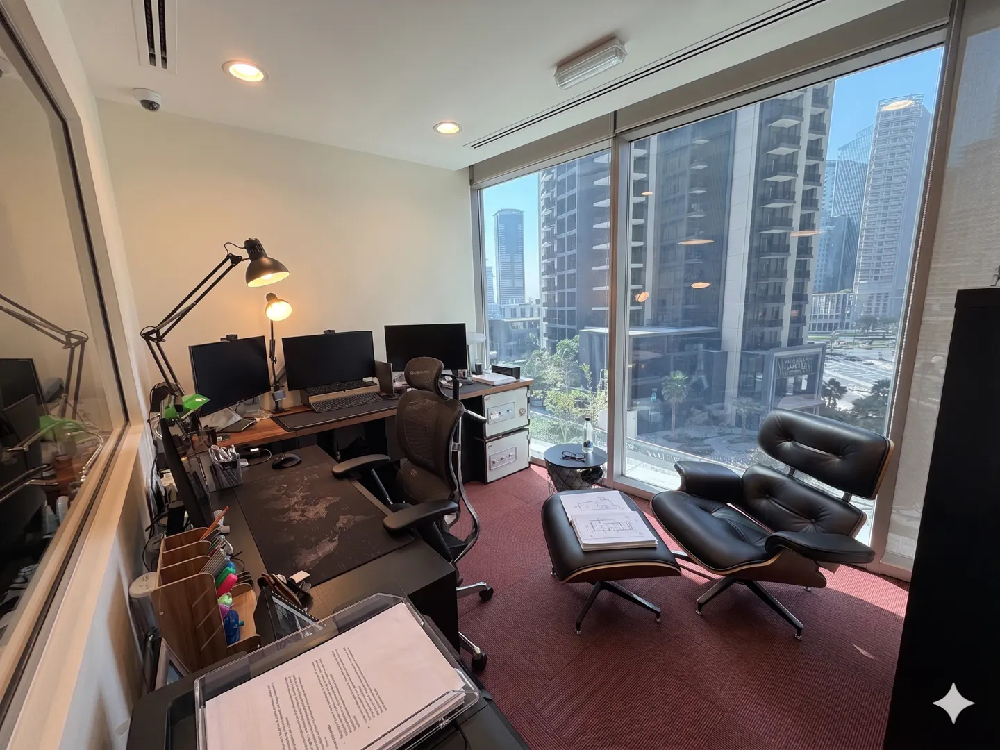 Office space with a desk, multiple monitors, and a black leather chair near a large window with a city view.