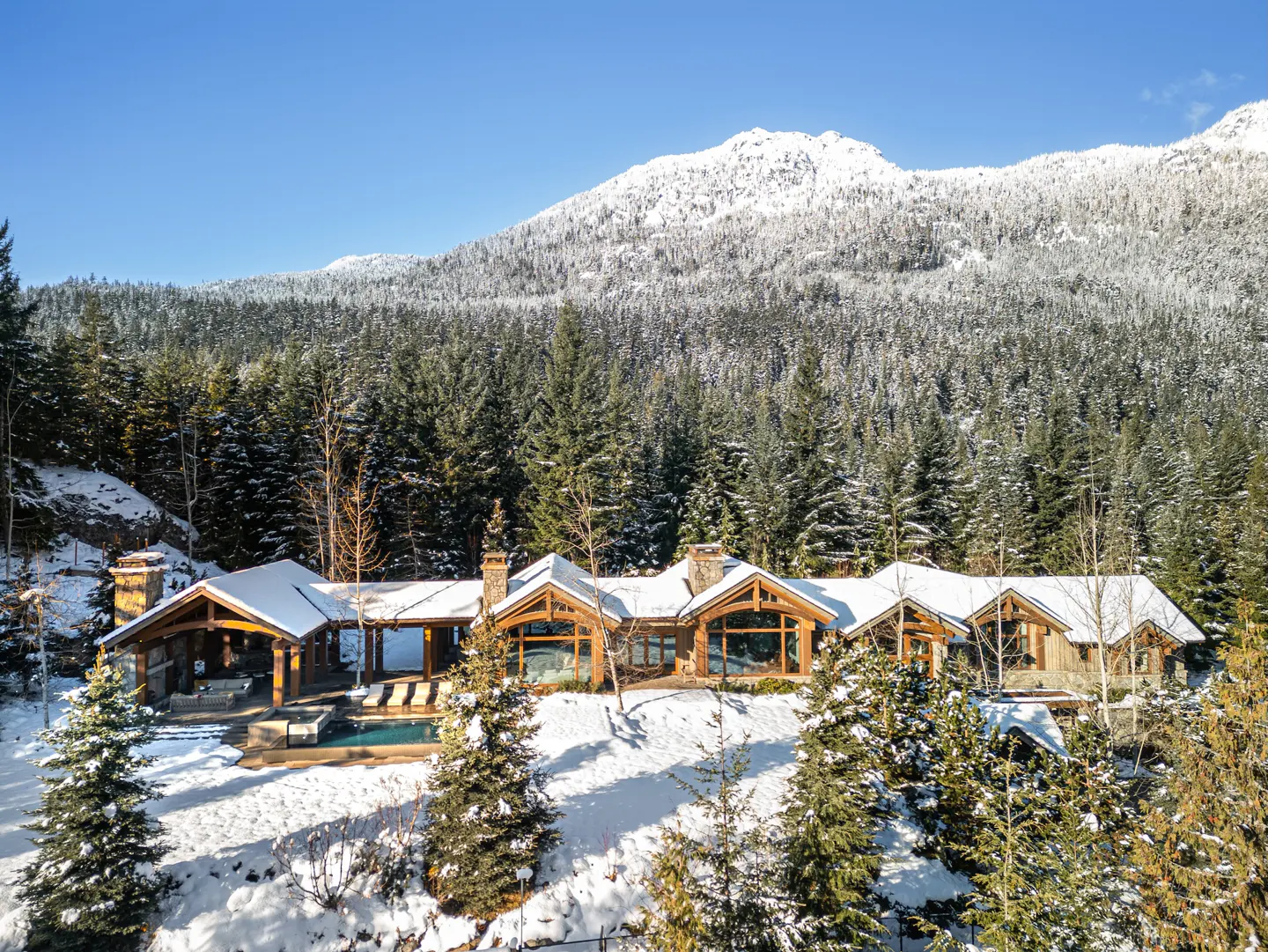 Luxury home with a pool in a snowy forest. The house has wood accents and a stone chimney. A snow-covered mountain is in the background.