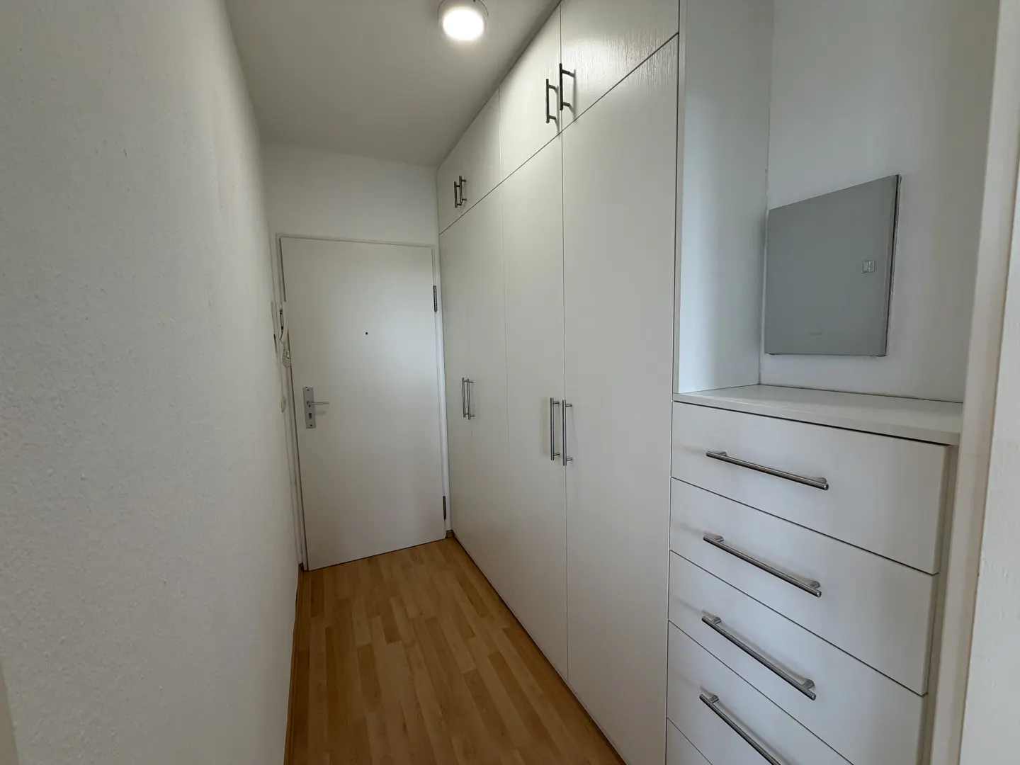 Hallway with light wood floors, white walls, and a white door. Built-in white cabinets and drawers line the right wall.