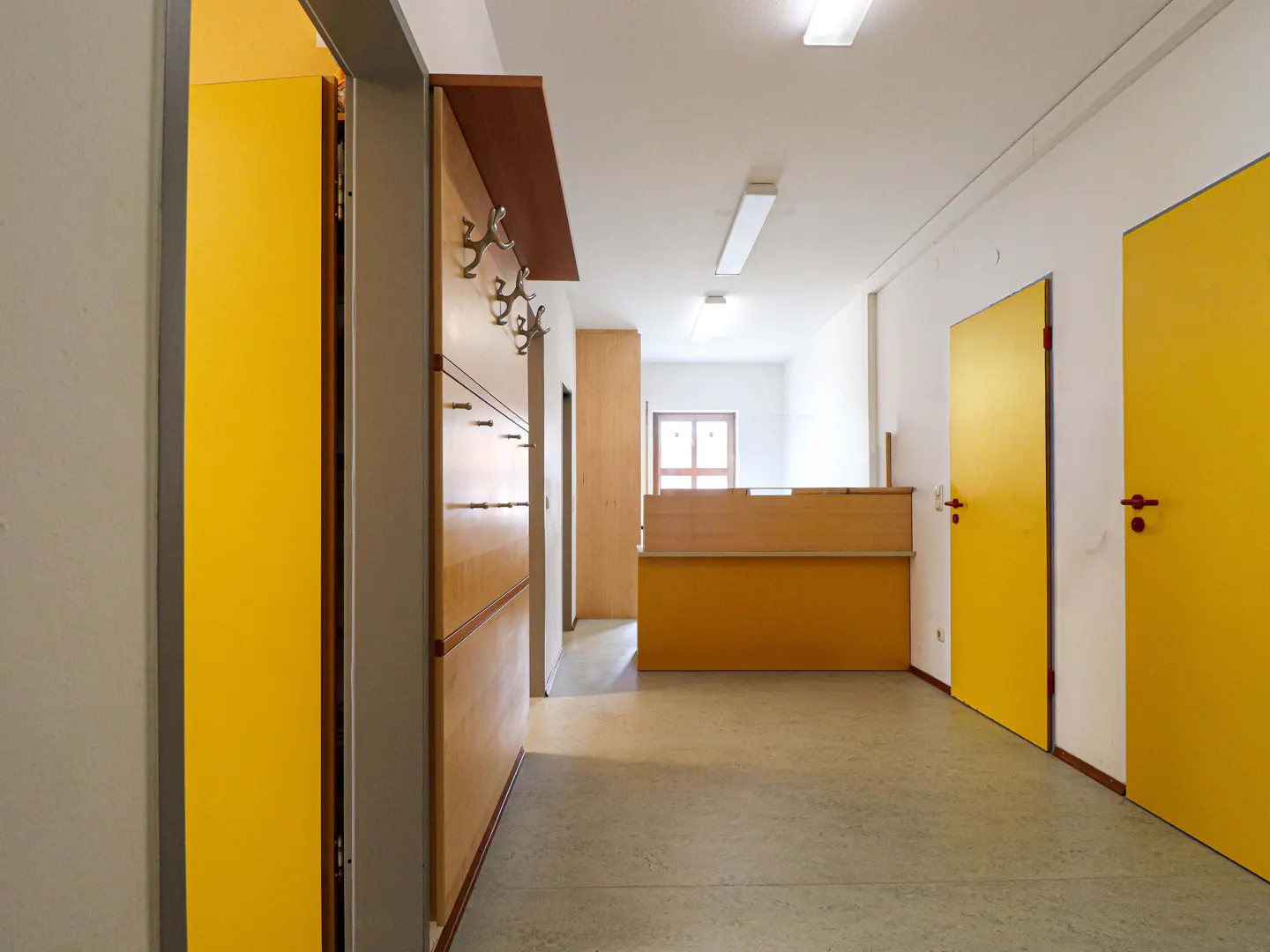 Hallway with yellow doors, coat rack, and reception desk. The walls are white and the floor is gray.