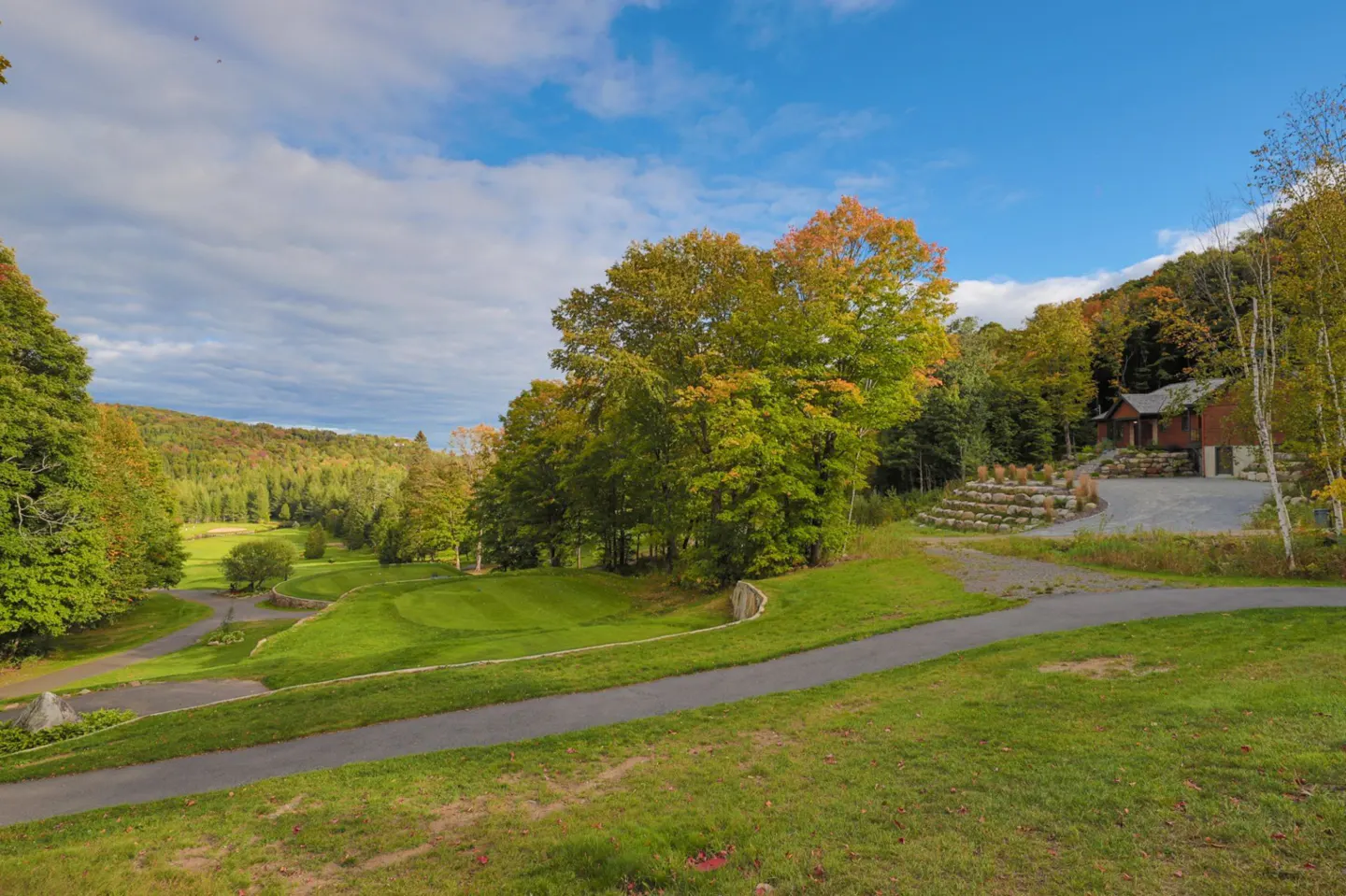 Scenic view of a golf course and a house nestled in the woods on a sunny day with blue skies and scattered clouds.