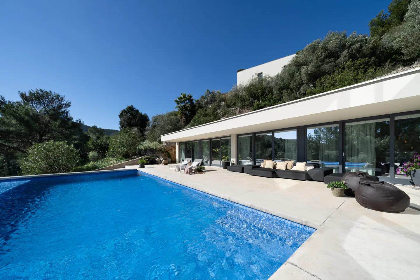 A modern white house with a blue tiled pool, a patio with black sofas, and a clear blue sky.