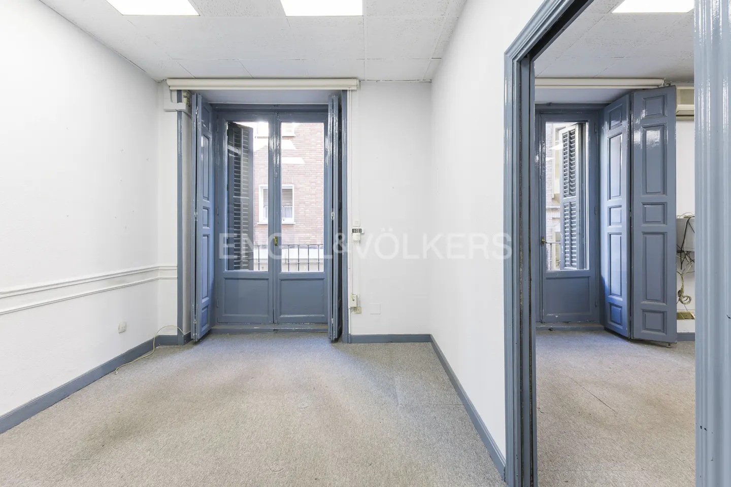 An empty room with gray carpet, white walls, and blue doors leading to a balcony and another room.