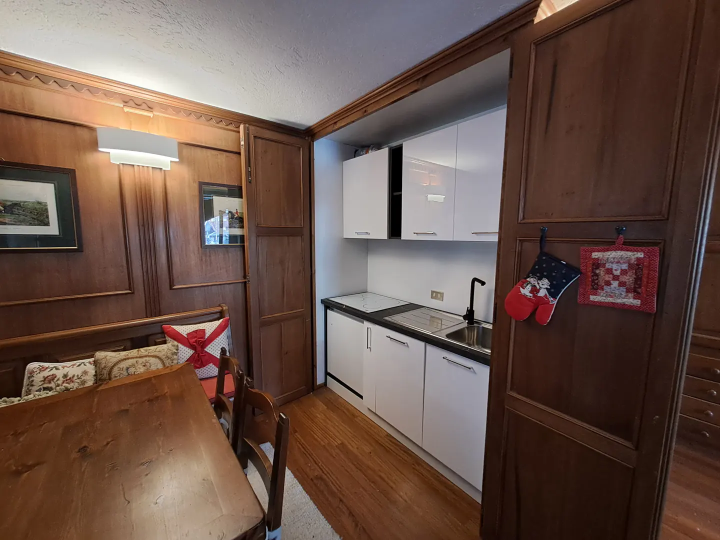 A cozy kitchen with wood paneling, a dining table, and white cabinets. Oven mitts hang on a wooden door.