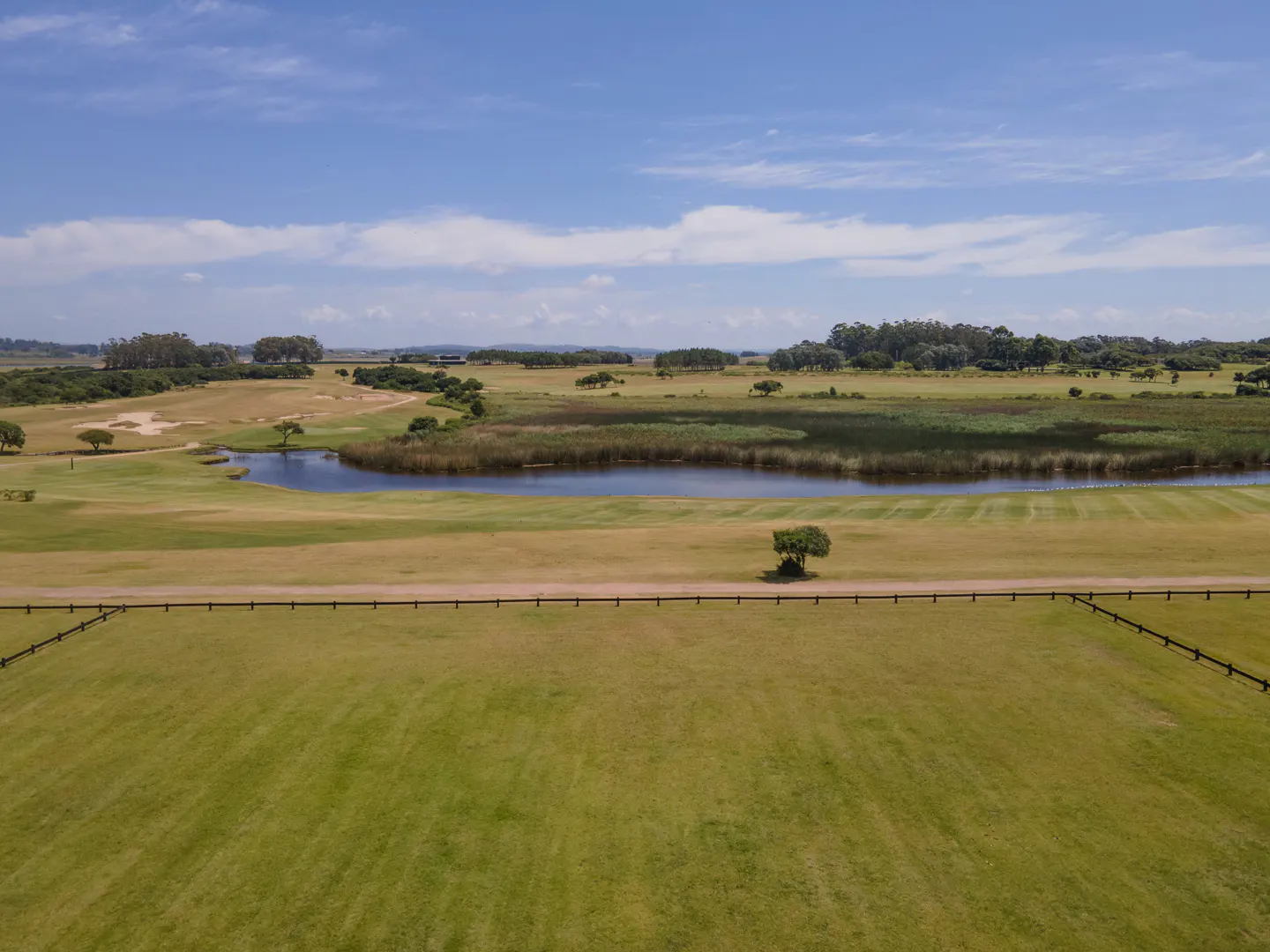 Aerial view of a green golf course with a pond, trees, and a wooden fence under a blue sky.