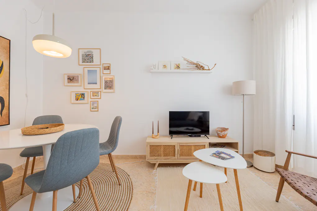 Bright living room with a dining area, blue chairs, a TV on a rattan console, and white nesting tables on a rug.