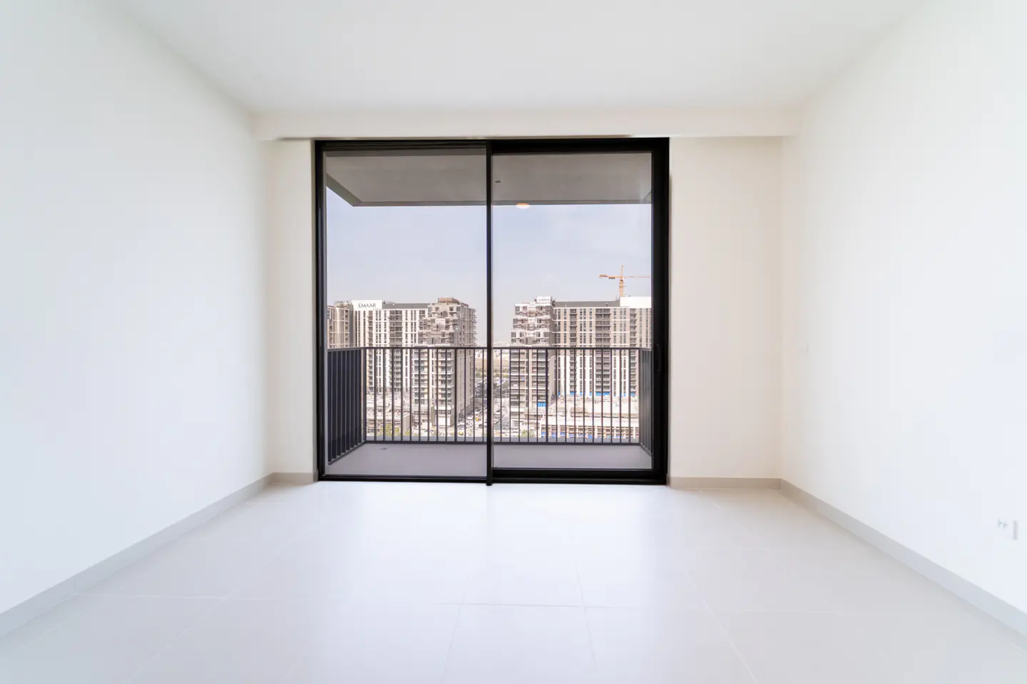 Empty white room with black-framed sliding glass doors leading to a balcony with city views.