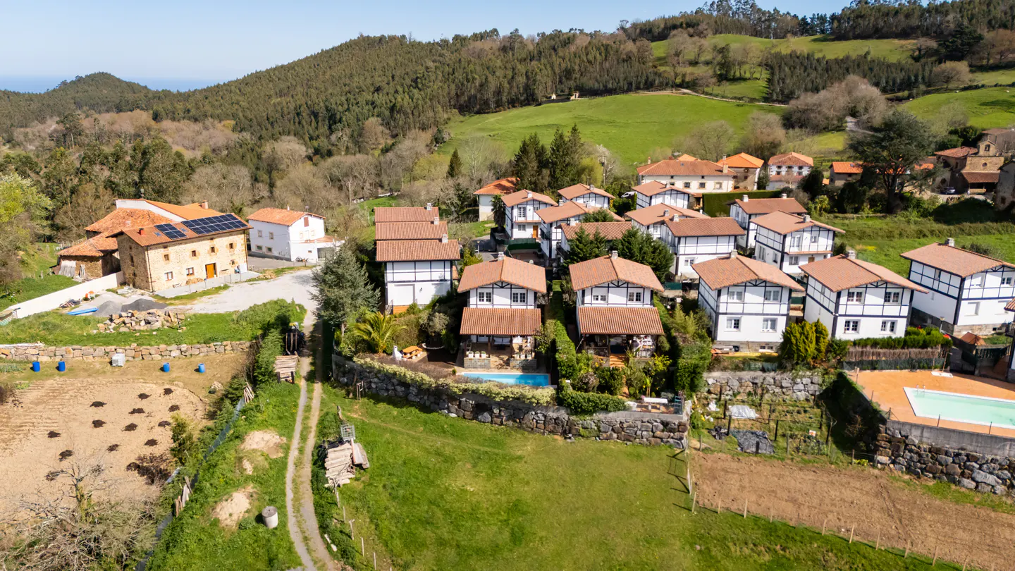 Aerial view of a rural village with white houses, red roofs, and green hills in the background. Some houses have pools.