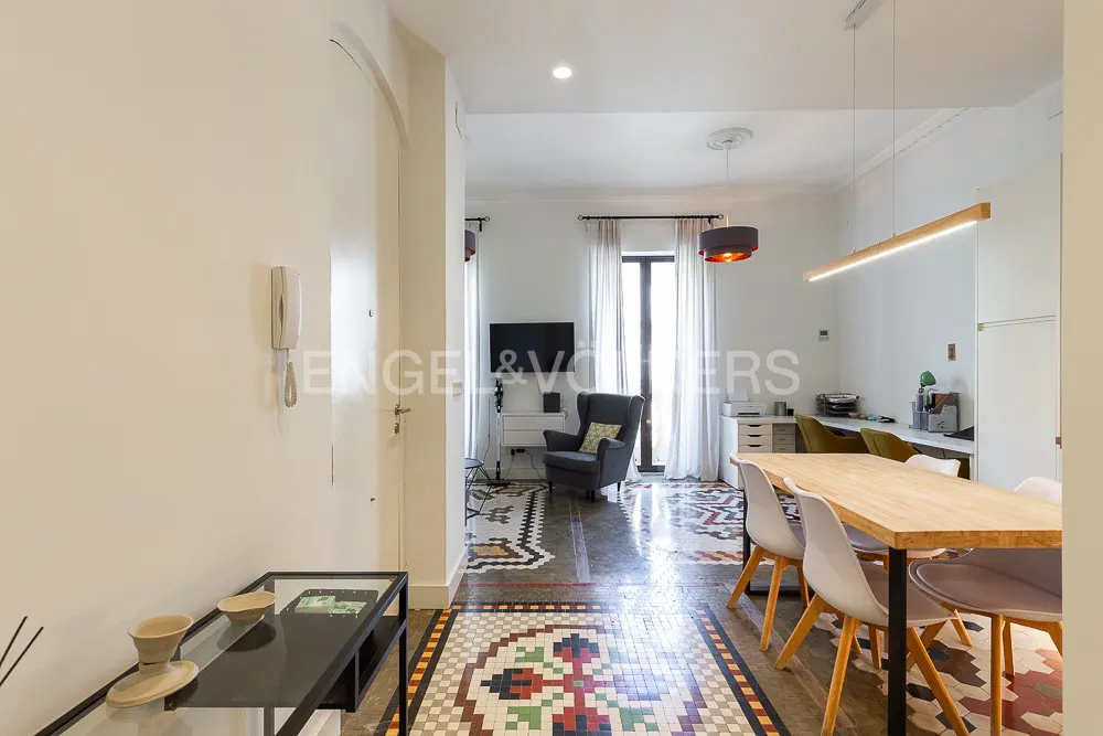 A bright, modern apartment interior with colorful tiled floors, a dining table with chairs, and a living area with a gray armchair.