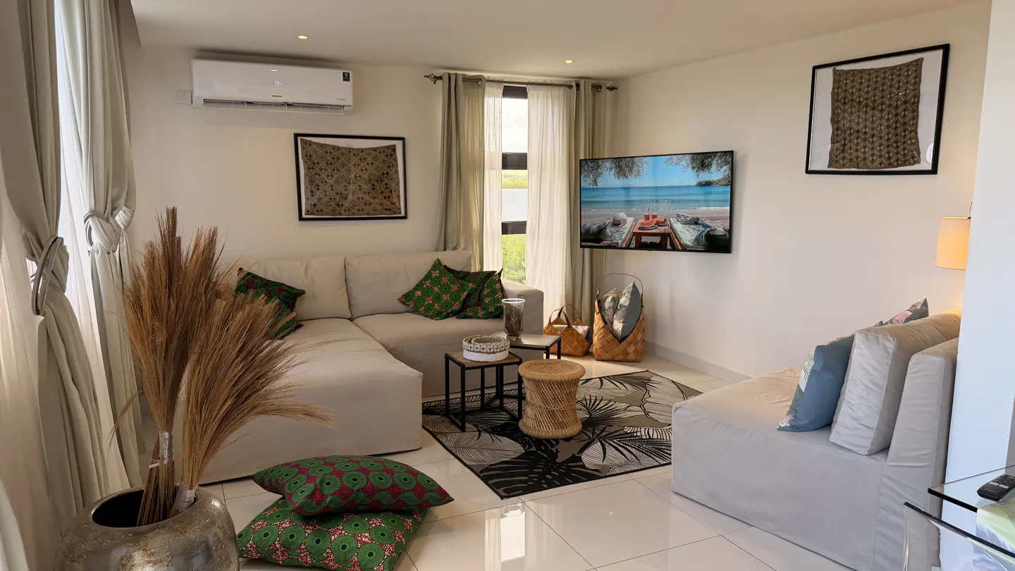 Living room with white walls, beige sofa, green patterned pillows, and a TV showing a beach scene. Artwork hangs on the walls.