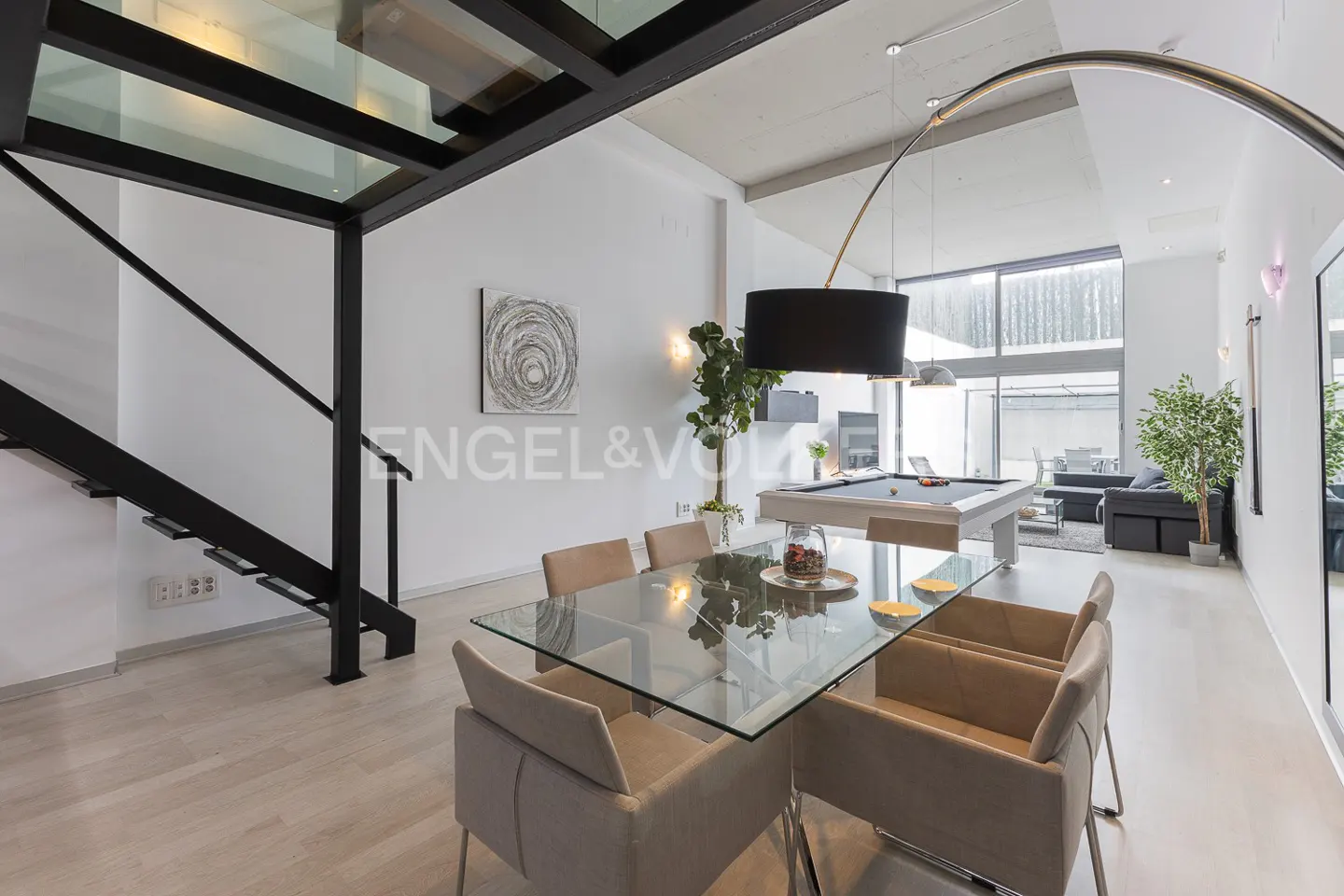 Bright, modern interior with glass table, beige chairs, and a pool table. Black metal staircase and large windows add architectural interest.