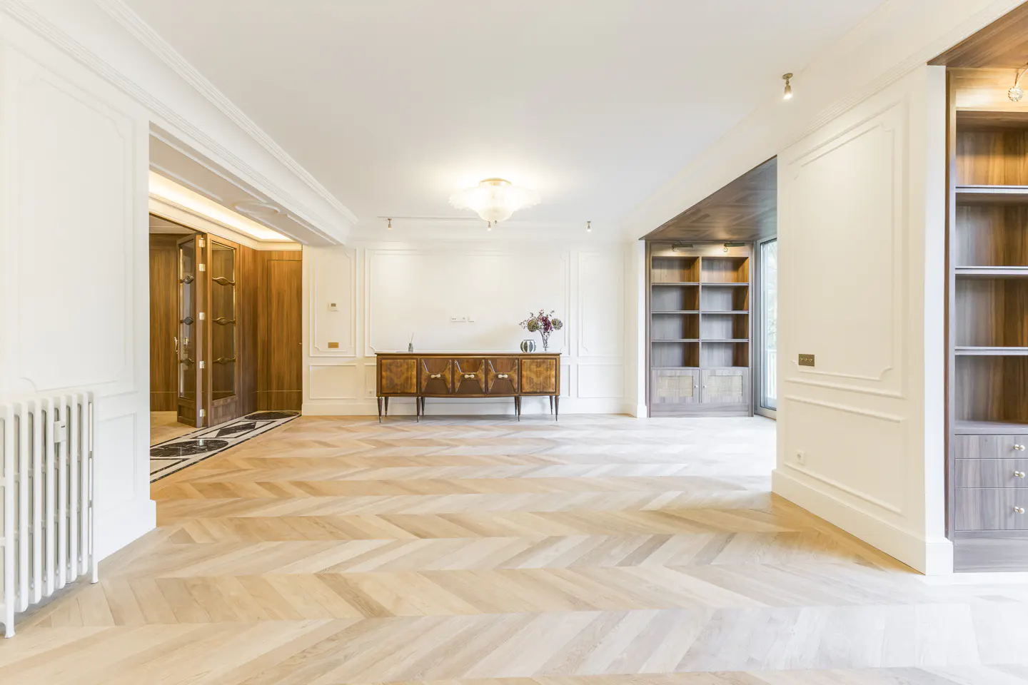 Bright, empty room with herringbone wood floors, white walls with molding, and a dark wood credenza with flowers. Built-in bookshelves are on either side of the room.