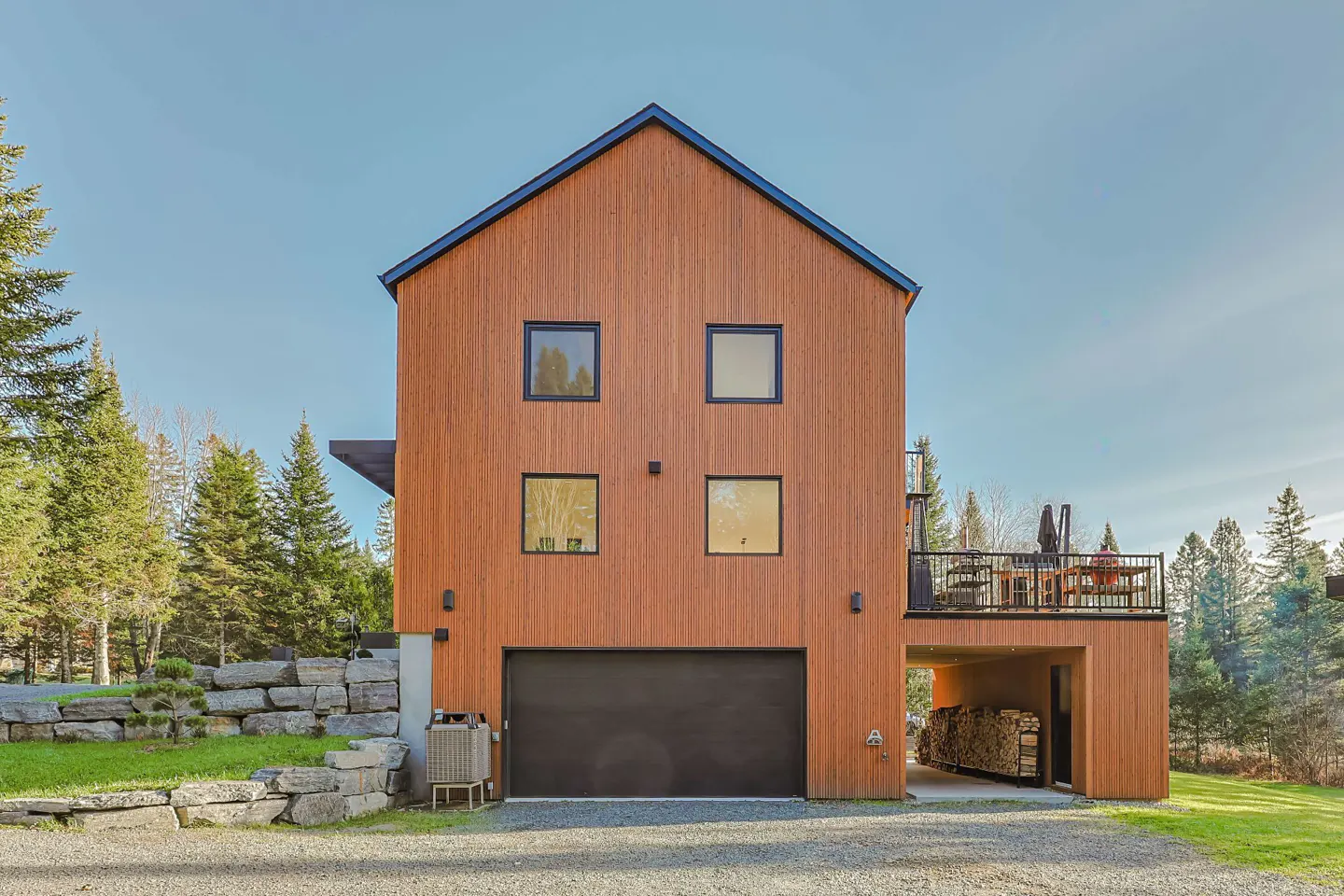 Modern wood-clad home with black trim, garage, and balcony. Trees and blue sky in the background.